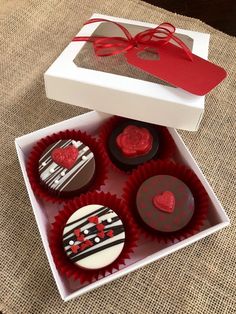 four decorated cupcakes in a white box with red ribbon and heart decorations on top