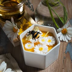 a bee sitting on top of a white bowl filled with liquid and daisies next to a jar of honey