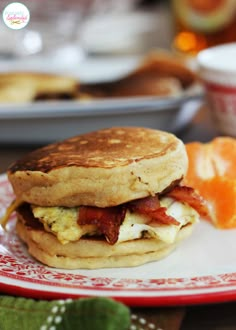 a breakfast sandwich with bacon and eggs on a red and white plate next to an orange slice