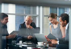 three people sitting at a table with laptops and papers in front of them, talking