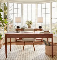 a wooden desk sitting in front of two large windows next to a potted plant