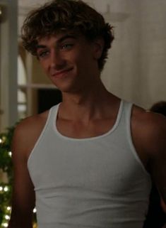 a young man standing in front of a christmas tree wearing a white tank top and looking at the camera