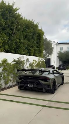 a grey sports car parked in front of a white house with trees and bushes behind it