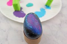 an egg sitting on top of a table next to a paper plate with colorful paint