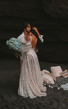 a woman in a white dress holding a bouquet of flowers and standing on the beach