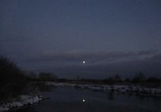 the moon is setting over a river with snow on the ground