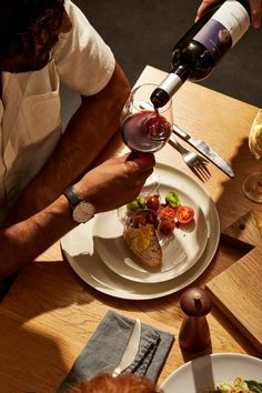 a person pouring wine into a glass on top of a plate with food and utensils