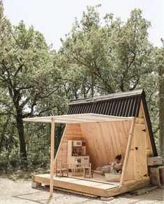 a small wooden cabin in the woods with a woman sitting on it's bed