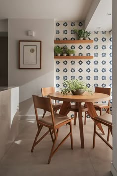 a dining room table and chairs with potted plants on the shelf above it, in front of a wallpapered background