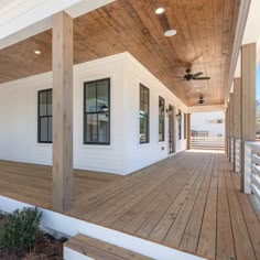 a porch with wooden flooring and ceiling fans on the side of it, next to a white house