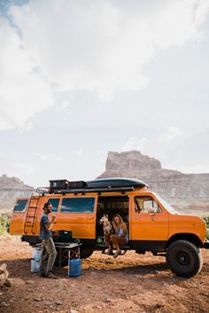 an orange truck parked in the desert with people sitting on it's bed and two dogs