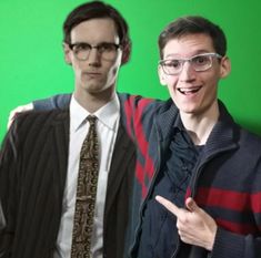 two men standing next to each other in front of a green screen with one pointing at the camera