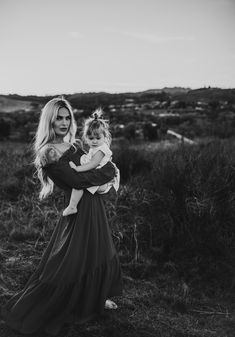 a woman holding a baby in her arms while standing in a field with grass and bushes