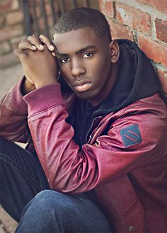 a young man leaning against a brick wall with his hands on his head and looking at the camera