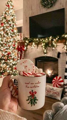 a person holding a coffee cup with marshmallows in front of a christmas tree