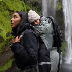 two people standing in front of a waterfall with backpacks on their backs and one is looking at the camera