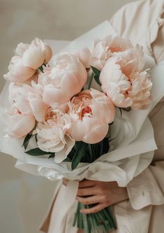 a woman holding a bouquet of pink peonies in her hands and wearing a white coat