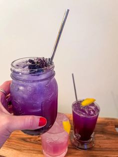 a person holding a purple drink in front of two other drinks on a wooden tray