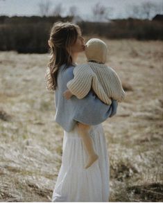 a woman holding a child in her arms while standing on a field with dry grass