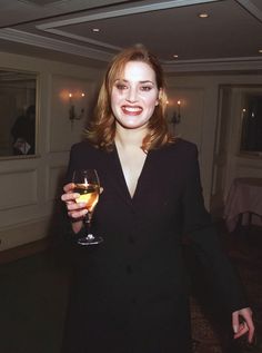 a woman in a black suit holding a glass of wine and smiling at the camera