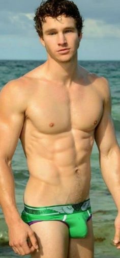 a shirtless man standing on the beach in his green swim trunks and no shirt