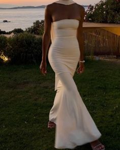 a woman in a white dress is walking on the grass near the ocean at sunset