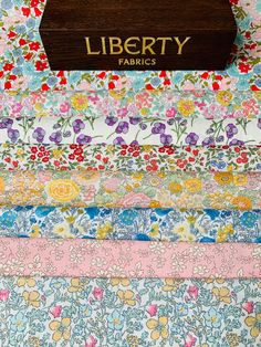 liberty fabrics is stacked on top of each other in different colors and patterns, with a wooden sign that reads liberty fabrics