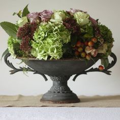 a vase filled with green and purple flowers on top of a white table cloth next to a wall