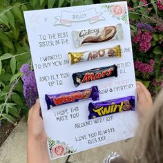 a person holding up a sign with candy on it and some flowers in the background