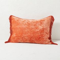 an orange velvet pillow with fringes on the edges, against a white wall background