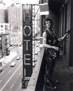 a man holding an electric guitar standing next to a hotel sign on the side of a road