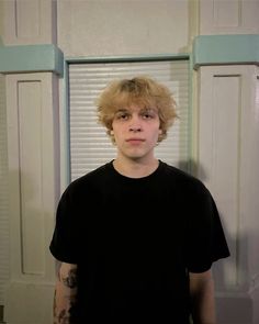 a young man with blonde hair standing in front of a door and looking at the camera