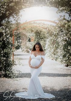 a pregnant woman in a white gown standing under an archway with flowers on the side