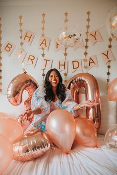 a woman sitting on top of a bed surrounded by pink balloons and gold foil letters