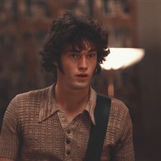 a young man with curly hair wearing a brown shirt and carrying a black bag looking at the camera