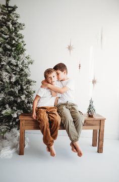 two young boys sitting on a bench in front of a christmas tree with their arms around each other