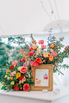 an arrangement of colorful flowers in a wooden frame on top of a white table under a tent