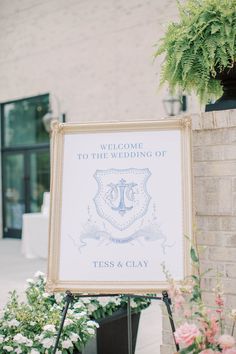 a welcome sign in front of a brick building with flowers and greenery around it