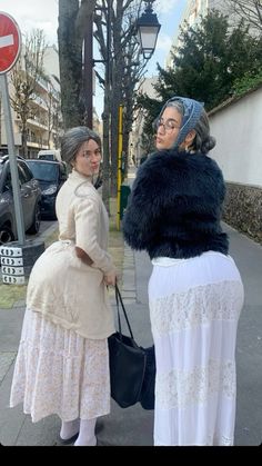 two women are standing on the sidewalk talking to each other and one is holding a purse