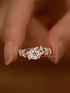 a woman's hand holding an engagement ring with leaves on the band and a diamond center stone