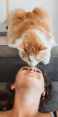 a woman laying on top of a couch next to an orange and white cat sitting on her head