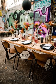 a wooden table topped with black plates and vases filled with flowers next to a graffiti covered wall