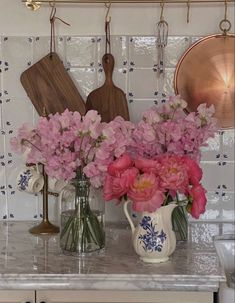 pink flowers are in vases on the kitchen counter next to utensils and cutting boards