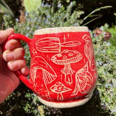 a hand holding a red and white coffee mug in front of some bushes with birds on it