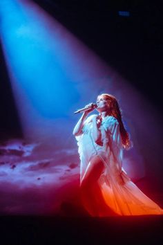 a woman standing on top of a stage with a microphone in her hand and lights behind her