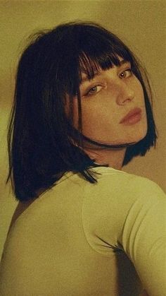 a woman with short hair wearing a white shirt and looking off to the side while standing in front of a wall