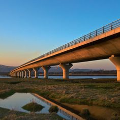 a bridge over a body of water with grass in the foreground and mountains in the background