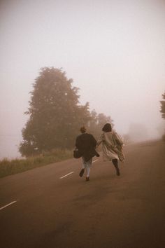 two people walking down the road on a foggy day with trees in the background