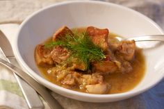 a white bowl filled with meat and gravy on top of a table next to a spoon