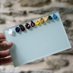 a hand holding a small white board with seven different colored beads on it's side